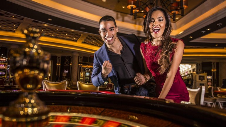 Молодой мужчина восточных кровей и его сексуальная спутница в обтягивающем бордовом платье эмоционально играют в рулетку в дорогом казино